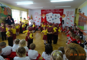 Na tle dekoracji z mapą Polski i dziećmi z chorągiewkami przedszkolaki tańczą w parach.